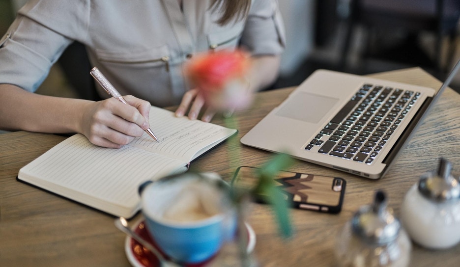 A desk with a laptop and a woman writing in the notebook with a pen