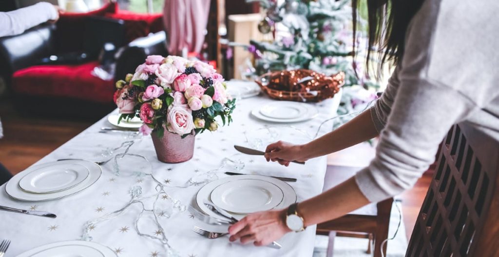 A woman setting the table with tablecloth, china plates, and a pink rose centerpiece.