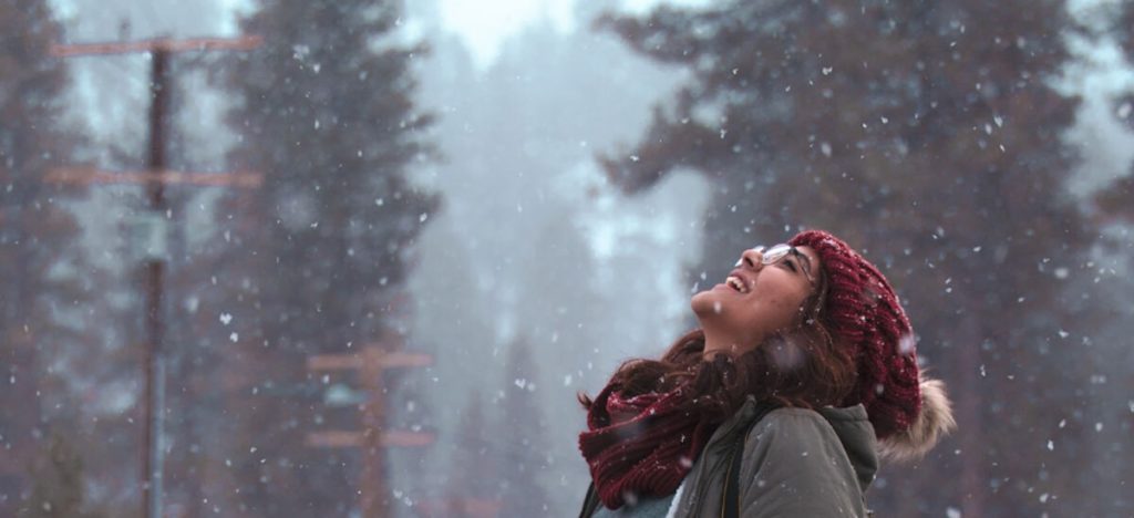A woman in a warm cap and scarf looking up into a snowing sky