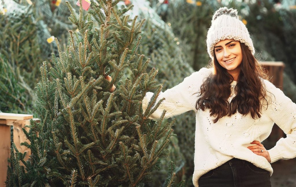girl in a warm hat and sweater standing next to a holiday tree