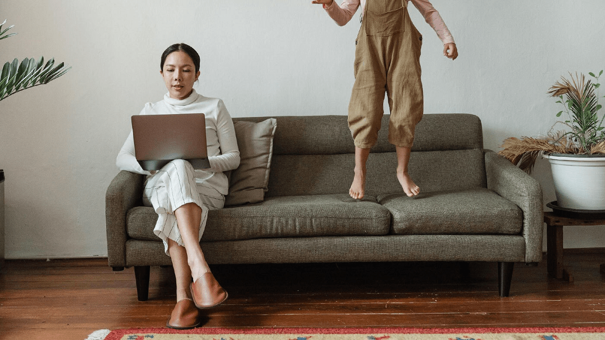 A woman sitting on the couch with a laptop and a child jumping on it next to her