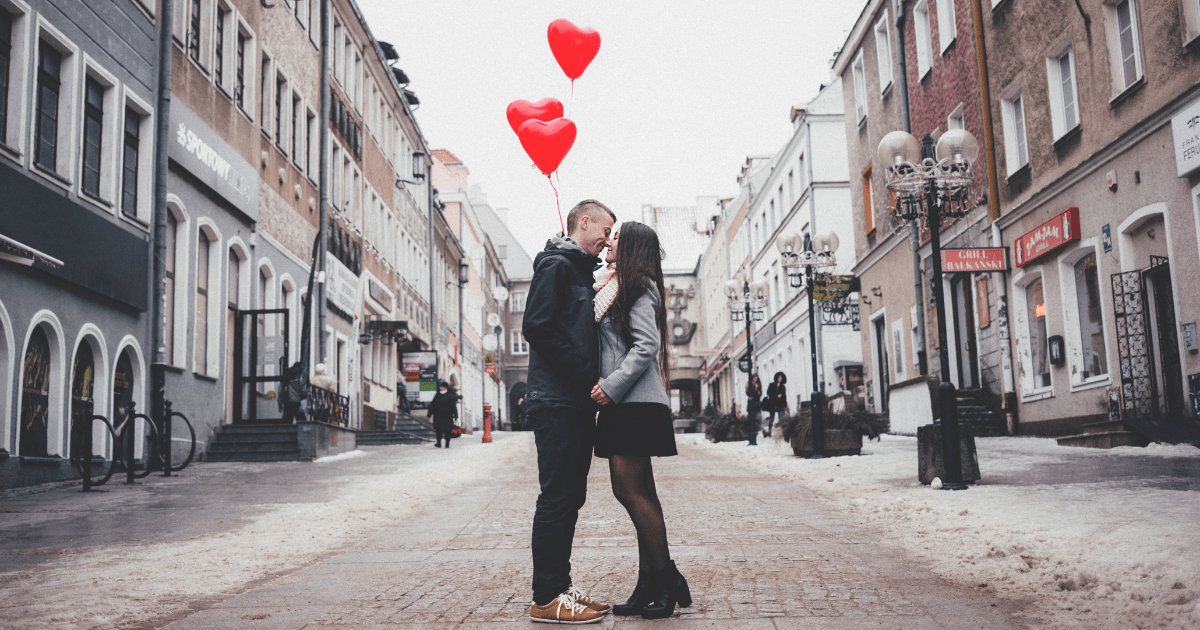 A couple facing each other in the middle of a city street holding red heart balloons