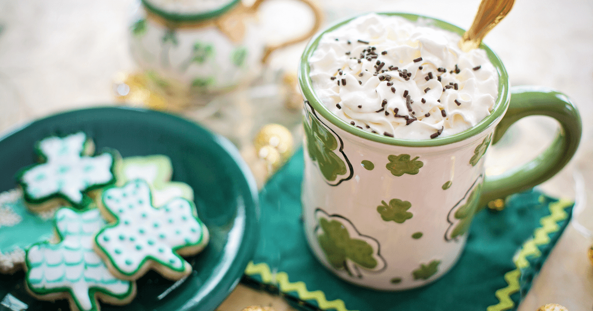 top down view of a plate of decorated cookies shaped as clovers and an Irish mug with whipped cream and sprinkles