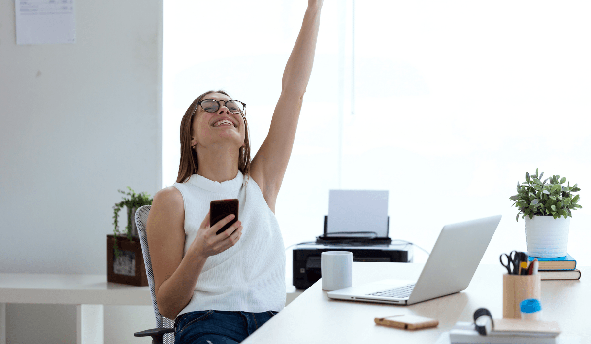 Lady at a desk with one arm up in the air and she's smiling, holding a phone and her laptop is open