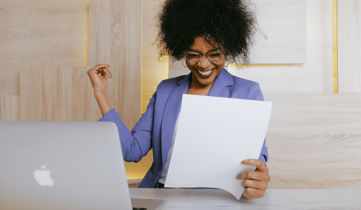 a person very happy and smiling while looking at a piece of paper next to her laptop