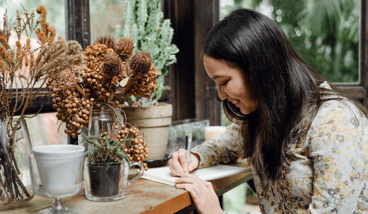 A woman at a table writing, surrounded by plants