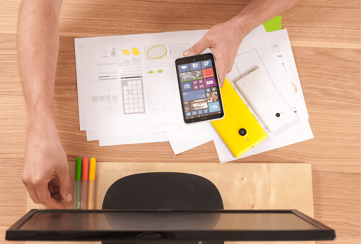 A top down view of a person holding their phone with work papers underneath on a desk and a desktop monitor