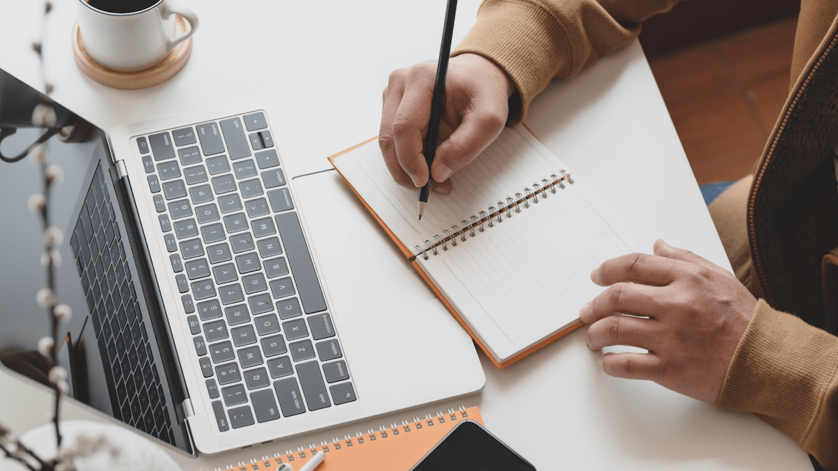A top down view of a hand writing in a journal on a desk next to a laptop